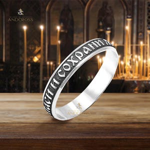 May include: A silver-toned metal ring featuring a band with engraved Cyrillic text in a dark, recessed finish. The ring has a smooth, polished interior surface and a textured outer band that displays the inscription clearly against the metal.