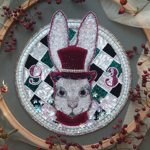 May include: A detailed, handmade brooch featuring a white rabbit with a top hat, set against a checkered background. The rabbit is adorned with beads and sequins, and the brooch is framed by a beaded border. The numbers "9" and "3" are also included.