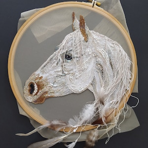 Puede incluir: Una cabeza de caballo blanca bordada con perlas y lentejuelas, enmarcada en un aro con un fondo de tela transparente. La crin del caballo está hecha de hilo blanco y plumas.