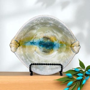 May include: A handcrafted ceramic bowl with a unique design. The bowl features a central splash of teal and brown, surrounded by yellow and white crystalline patterns. The rim has small handles. It is displayed on a black metal stand.