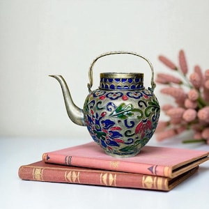 May include: A decorative, vintage teapot with a silver spout and handle. The body is adorned with a colorful floral design in red, blue, and green. The teapot sits atop two antique books with pink covers.