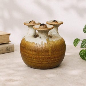 May include: A round ceramic vase with three distinct spouts, each topped with a small spherical stopper. The base features a deep brown glaze that transitions into a lighter, crackled cream finish toward the top of the three neck openings.