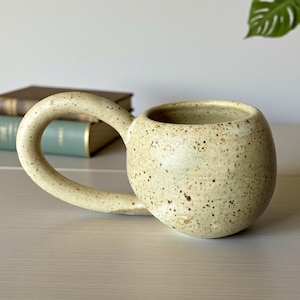 May include: A white ceramic mug with a black handle and the text "But First, Coffee" in a bold, black font. The mug is set against a blurred background, suggesting a cozy setting. The mug appears to be approximately 4 inches tall.