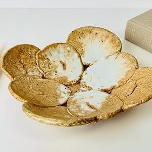 May include: A handcrafted ceramic bowl with a unique, organic design. The bowl features a series of overlapping, petal-like shapes in shades of brown and white, with a textured surface. The rim is accented with a gold-colored glaze. A decorative piece for the home.