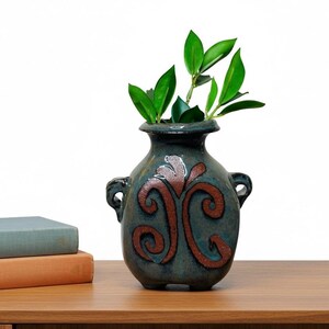 May include: A teal ceramic vase with brown swirl designs and two small handles. The vase holds green leafy stems. The vase sits on a wooden surface, with two books visible in the background.