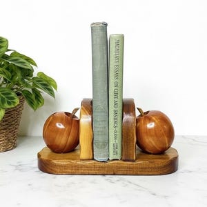 May include: Wooden apple-shaped bookends with a natural finish. The bookends hold two vintage books with green covers. The base is a wooden platform. A potted plant is in the background.