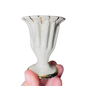 K&ouml;nnte beinhalten: Kleine, wei&szlig;e Keramikvase mit geriffeltem Design und Goldrand. Die Vase hat einen Sockel und eine ausgestellte Oberseite. Die Vase wird in einer Hand vor wei&szlig;em Hintergrund gehalten. Wahrscheinlich Vintage oder Antik.