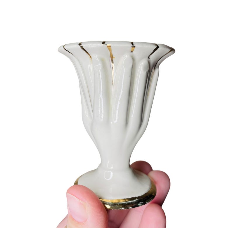 K&ouml;nnte beinhalten: Kleine, wei&szlig;e Keramikvase mit geriffeltem Design und Goldrand. Die Vase hat einen Sockel und eine ausgestellte Oberseite. Die Vase wird in einer Hand vor wei&szlig;em Hintergrund gehalten. Wahrscheinlich Vintage oder Antik.