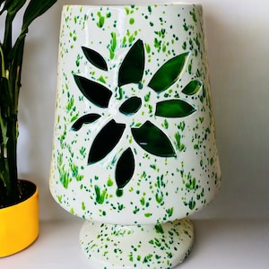 May include: White ceramic vase with a green floral design. The vase has a wide base and a narrow neck. The floral design is cut out of the vase, creating a decorative effect.