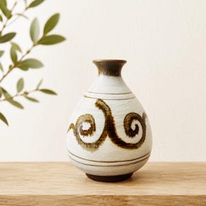 May include: A ceramic vase with a rounded body and a narrow neck, featuring a cream-colored glaze with hand-painted dark brown swirling patterns and horizontal lines. The rim and base are finished in a solid dark brown tone.