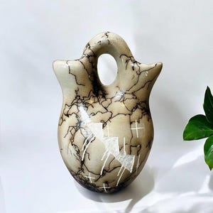 May include: A hand-painted ceramic Native American pottery vase with a light brown background and black and white designs. The vase has a unique shape with a rounded body and a narrow neck.
