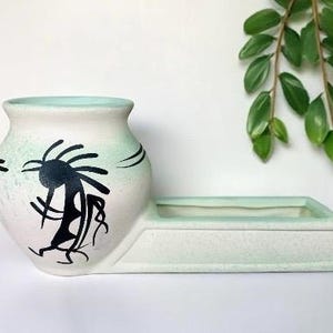 May include: A ceramic planter in white and mint green. The vase has a black Kokopelli design. The planter has a rectangular base attached to the vase. Green leaves hang in the background.