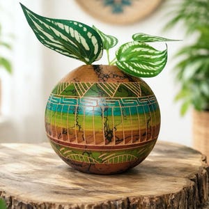 May include: A round, hand-painted planter with a vibrant, geometric design in shades of green, blue, and brown. The planter holds a small plant with green and white striped leaves. The planter is displayed on a wooden tree stump.