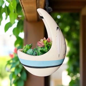 May include: Cream-colored hanging planter with a blue stripe and black accents, filled with colorful succulents. The planter has a unique crescent shape with a feather design. The planter is suspended from a black rope.