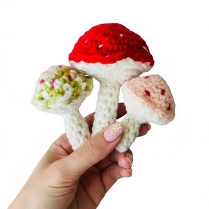 May include: Three small crocheted mushroom figures with white stems. The central mushroom has a bright red cap with white spots. The left mushroom features a textured green and white cap, while the right mushroom has a soft peach-colored cap.