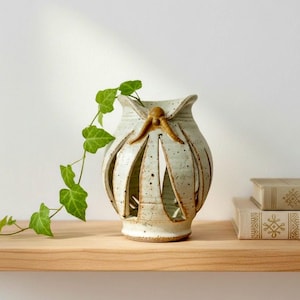May include: A handmade ceramic vase with a cream-colored glaze and cut-out design. The vase has a brown accent and is decorated with small brown speckles. A green vine with leaves is draped over the vase. The vase is on a wooden shelf.