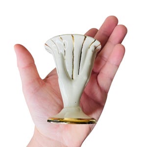 K&ouml;nnte beinhalten: Kleine, cremefarbene Keramikvase in Form einer Hand, mit goldenen Akzenten. Die Vase hat eine ausgestellte Oberseite und einen Sockel, die beide mit Gold verziert sind. Die handf&ouml;rmige Vase wird in der Hand einer Person gehalten.