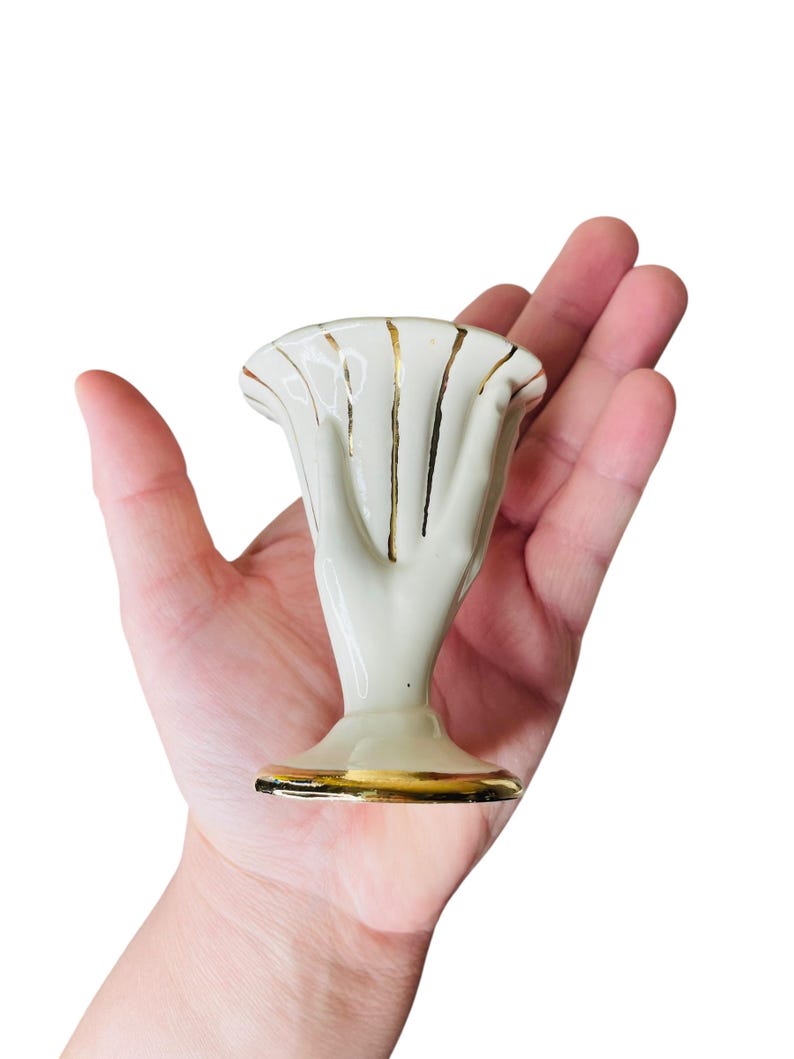 K&ouml;nnte beinhalten: Kleine, cremefarbene Keramikvase in Form einer Hand, mit goldenen Akzenten. Die Vase hat eine ausgestellte Oberseite und einen Sockel, die beide mit Gold verziert sind. Die handf&ouml;rmige Vase wird in der Hand einer Person gehalten.