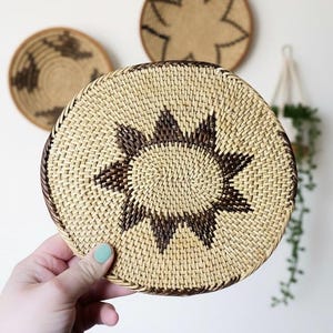 May include: A round, woven basket with a sunburst design in shades of brown and tan. The basket is held in a hand, with two other decorative baskets in the background. The baskets are likely for wall decor.