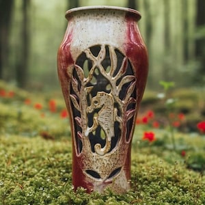 May include: A handcrafted ceramic vase with a seahorse and tree design. The vase features a red and cream color scheme with cut-out details. The vase is cylindrical with a wide opening and is set against a blurred forest background.