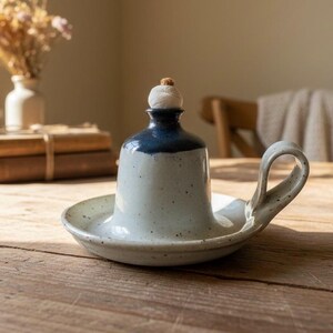 May include: A handmade ceramic oil lamp with a light blue and dark blue glaze. The lamp features a small handle and a wick. The lamp is sitting on a wooden table. The lamp is a decorative item.