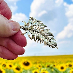 May include: A gold-toned, leaf-shaped brooch with clear crystal accents. The brooch is held against a backdrop of a bright blue sky with fluffy white clouds and a field of yellow sunflowers.
