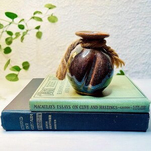 May include: A small, dark brown and blue ceramic vase with a rope handle sits on top of two books. The top book is green and has the title "Macaulay's Essays on Clive and Hastings" printed on the cover. The bottom book is blue and has the title "Chinese Curtain" printed on the cover.
