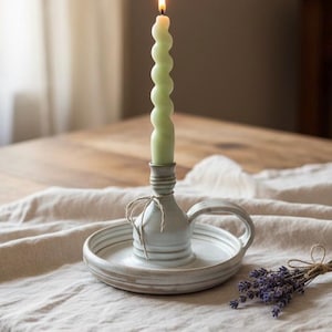 May include: A lit, light green, spiral-shaped candle sits in a ceramic candle holder with a handle and a round base. The candle holder is tied with twine. A small bundle of lavender is on the table.