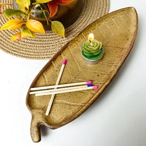 May include: A leaf-shaped ceramic tray in a speckled brown color holds a green rose-shaped candle and three matches with pink and blue tips. The tray has a textured surface and a small handle. A decorative plant is in the background.