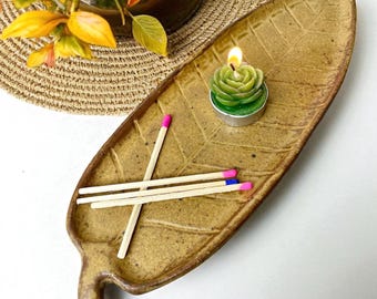 May include: A leaf-shaped ceramic tray in a speckled brown color holds a green rose-shaped candle and three matches with pink and blue tips. The tray has a textured surface and a small handle. A decorative plant is in the background.