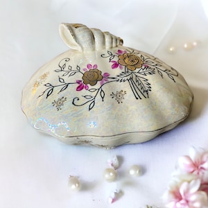 May include: A white ceramic trinket box with a floral design in gold and pink. The box has a shell-like shape with a decorative handle.