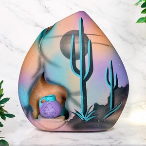 May include: A ceramic sculpture of a desert scene with a purple clay pot, two cacti, and a black sun. The sculpture is painted in shades of blue, pink, and brown.