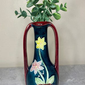 May include: A small, dark blue ceramic vase with red handles and floral designs in yellow, pink, and green. The vase is filled with a sprig of green eucalyptus.