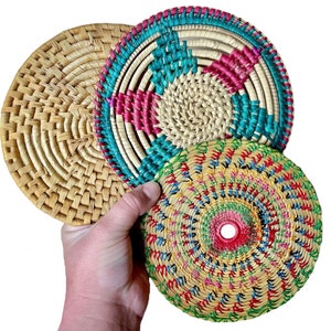 May include: Three woven round coasters with colorful patterns. The top coaster has a pink, blue, and green pattern. The middle coaster is a natural woven color. The bottom coaster has a multi-colored pattern with pink, yellow, green, and blue.