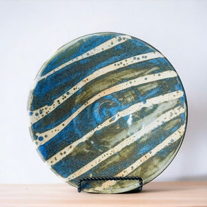 K&ouml;nnte beinhalten: Ein gro&szlig;er, runder Teller aus Keramik mit einem gestreiften Design in Blau, Gr&uuml;n und Wei&szlig;. Der Teller hat eine leichte Spiralform in der Mitte.