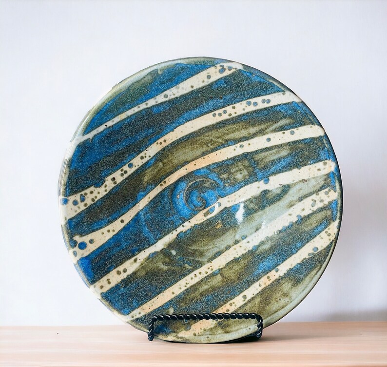 K&ouml;nnte beinhalten: Ein gro&szlig;er, runder Teller aus Keramik mit einem gestreiften Design in Blau, Gr&uuml;n und Wei&szlig;. Der Teller hat eine leichte Spiralform in der Mitte.