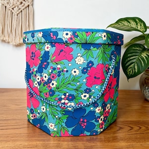 May include: A hexagonal floral storage box with a blue lid and handle. The box features a vibrant pattern of pink, blue, and white flowers on a turquoise background. A blue and white rope handle adds a decorative touch.