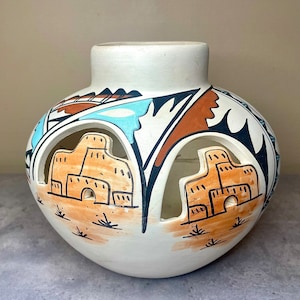 Peut inclure: Un pot en céramique blanche avec des motifs découpés et des détails peints à la main en brun, orange, bleu et noir. Le pot présente un design d'inspiration sud-ouest avec des motifs géométriques et des représentations stylisées de bâtiments.