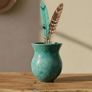 May include: A turquoise ceramic vase with a crackled glaze finish, featuring a rounded body and a flared rim. Two feathers are placed inside the vase: one is a vibrant turquoise with black markings, and the other is brown with white stripes.