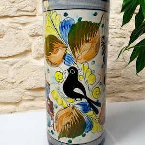 May include: A white ceramic vase with a black bird and colorful floral design. The vase has a cylindrical shape and is decorated with yellow, blue, green, and brown leaves.