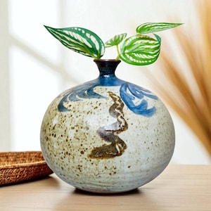 May include: A round ceramic vase with a speckled, off-white body and blue and brown painted details. The vase holds green and white striped leaves. The vase is sitting on a wooden surface.