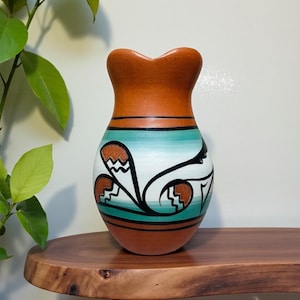 May include: A handcrafted ceramic vase with a unique shape and design. The vase features a brown top and base, with a turquoise and white painted body. Black and brown abstract patterns adorn the body, adding to its artistic appeal. The vase is a decorative item.