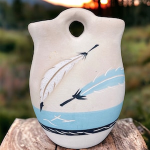 May include: A white ceramic vase with a blue and black design featuring two stylized feathers. The vase has a hole at the top for hanging.