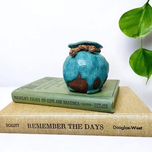 May include: A small, teal blue ceramic vase with a brown glaze and a leather strap around the top. The vase is sitting on top of two books.