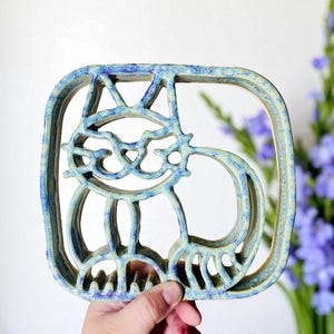 May include: A ceramic cat-shaped trivet with a blue and cream glaze. The cat design is open, with cut-out details forming the cat's features and body within a square frame. The trivet is held in a hand.