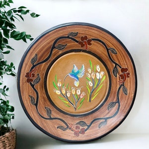 May include: A round wooden wall plate with a brown and black woodgrain finish. The plate features a painted design of a hummingbird flying over a branch with white flowers.