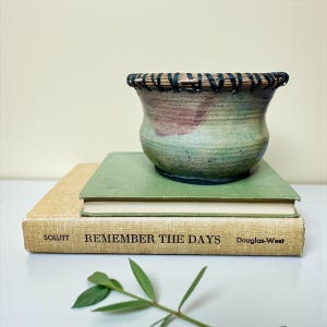 May include: A handcrafted ceramic bowl with a woven rim, resting on two vintage books. The bowl features a blend of green, pink, and brown hues. The books are a light green and tan color, with the text "REMEMBER THE DAYS" visible.