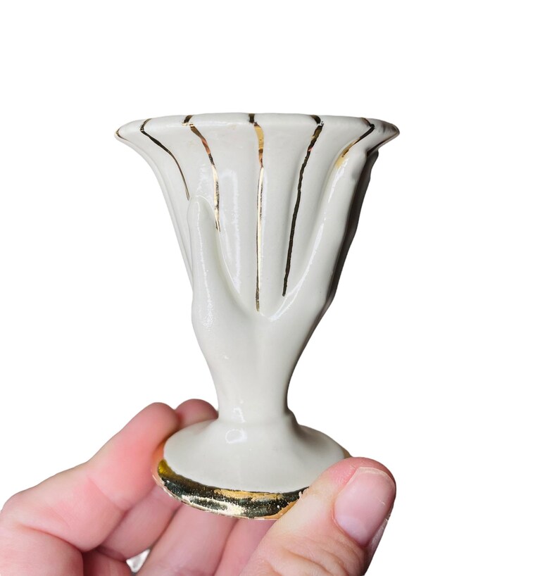 K&ouml;nnte beinhalten: Kleine, cremefarbene Keramikvase mit ausgestelltem Oberteil und Sockel. Die Vase hat vertikale Goldstreifen und Goldbesatz um den Boden und den Rand. Das Design ist elegant und vintage.