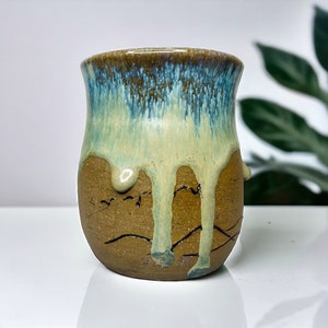 May include: A brown ceramic mug with a light blue and green glaze that drips down the sides. The glaze has a speckled appearance.