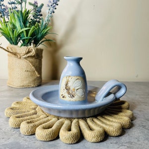 May include: A blue ceramic candle holder with a floral design. The holder is on a woven rope coaster.